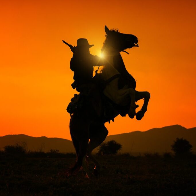 Silhouette cowboy holding short gun and riding a horse on sunrise