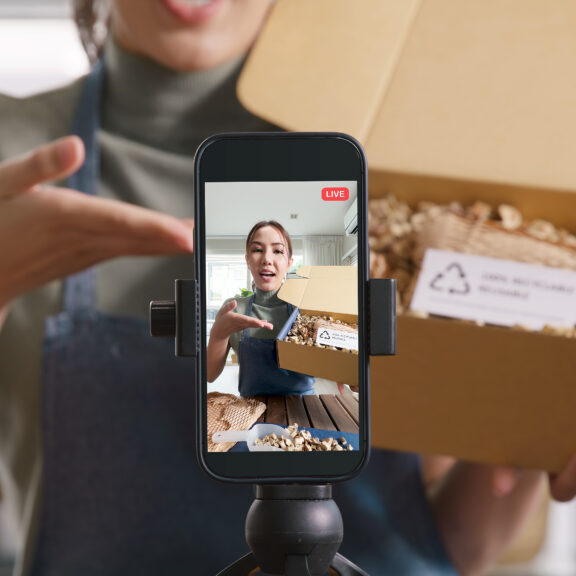 SME owner go green net zero retail store live stream talk on phone showing eco care plastic free packaging box. Asia people young woman record video on tiktok IG reel instagram shop happy side hustle.