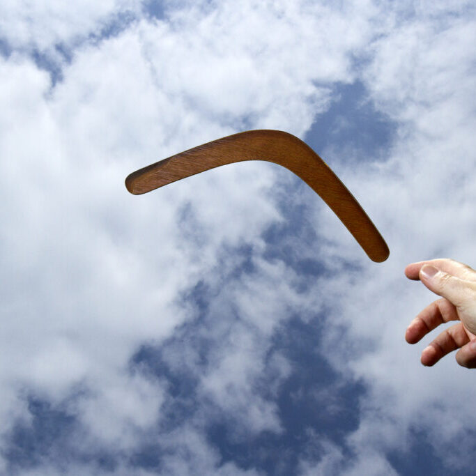 Throwing a plain wooden boomerang midair with blue sky and cloud background.