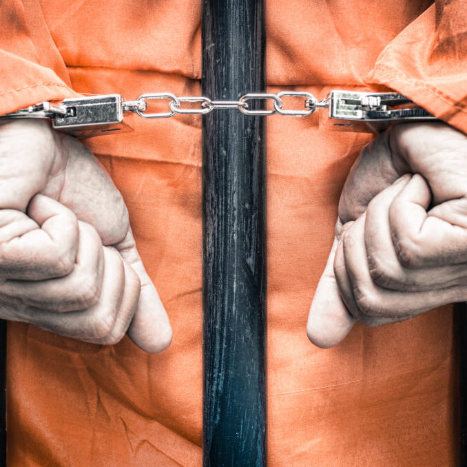 Handcuffed hands of a prisoner behind the bars of a prison with orange clothes - Crispy desaturated dramatic filtered look