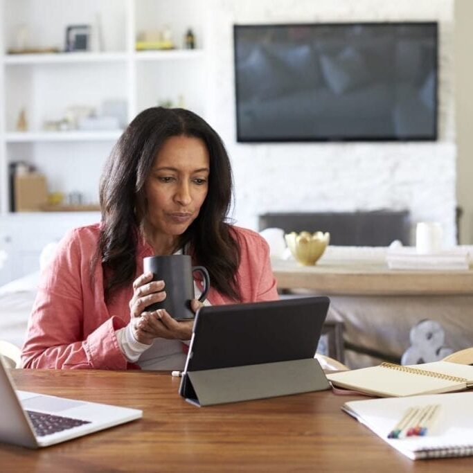 Middle aged woman sitting at a table reading using a tablet computer, holding a cup, front view