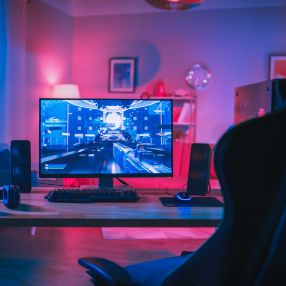 Powerful Personal Computer Gamer Rig with First-Person Shooter Game on Screen. Monitor Stands on the Table at Home. Cozy Room with Modern Design is Lit with Pink Neon Light.