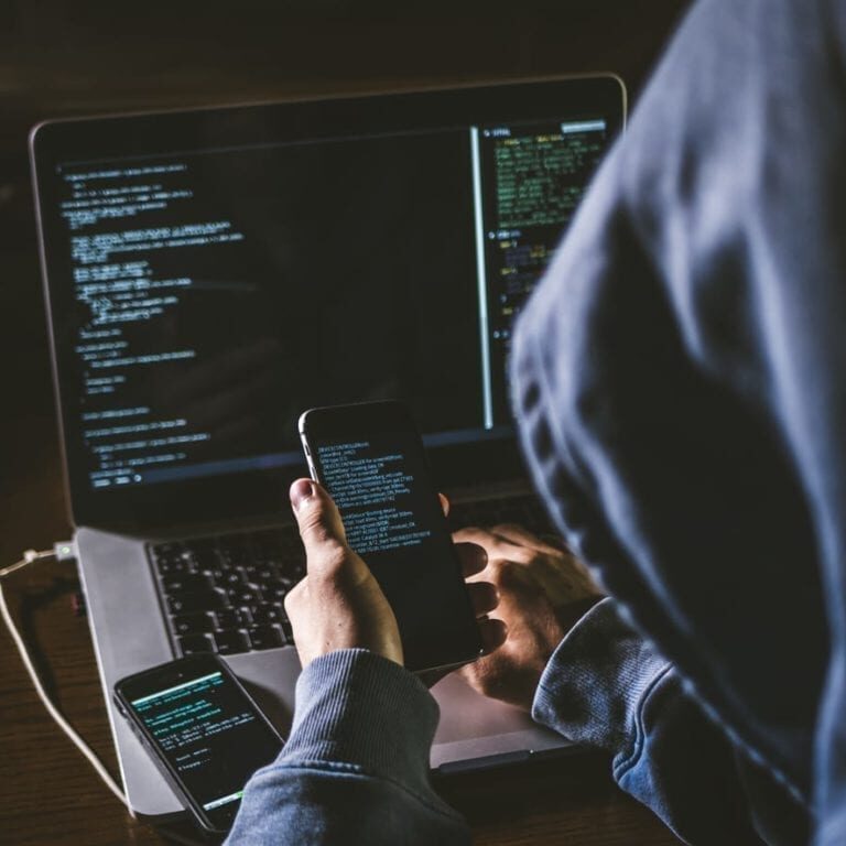 anonymous person in the hood sitting in front of computer working with laptop and mobile phone