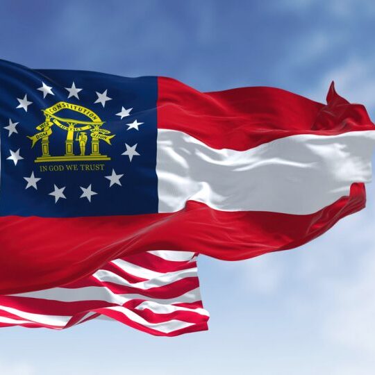 The Georgia state flag waving along with the national flag of the United States of America. In the background there is a clear sky.