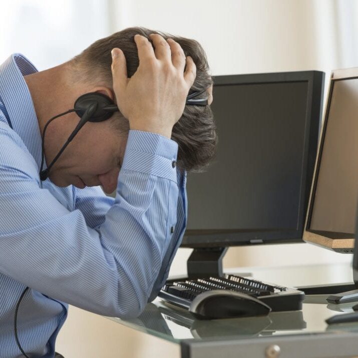Profile shot of male exhausted trader with head in hands leaning at computer desk in office